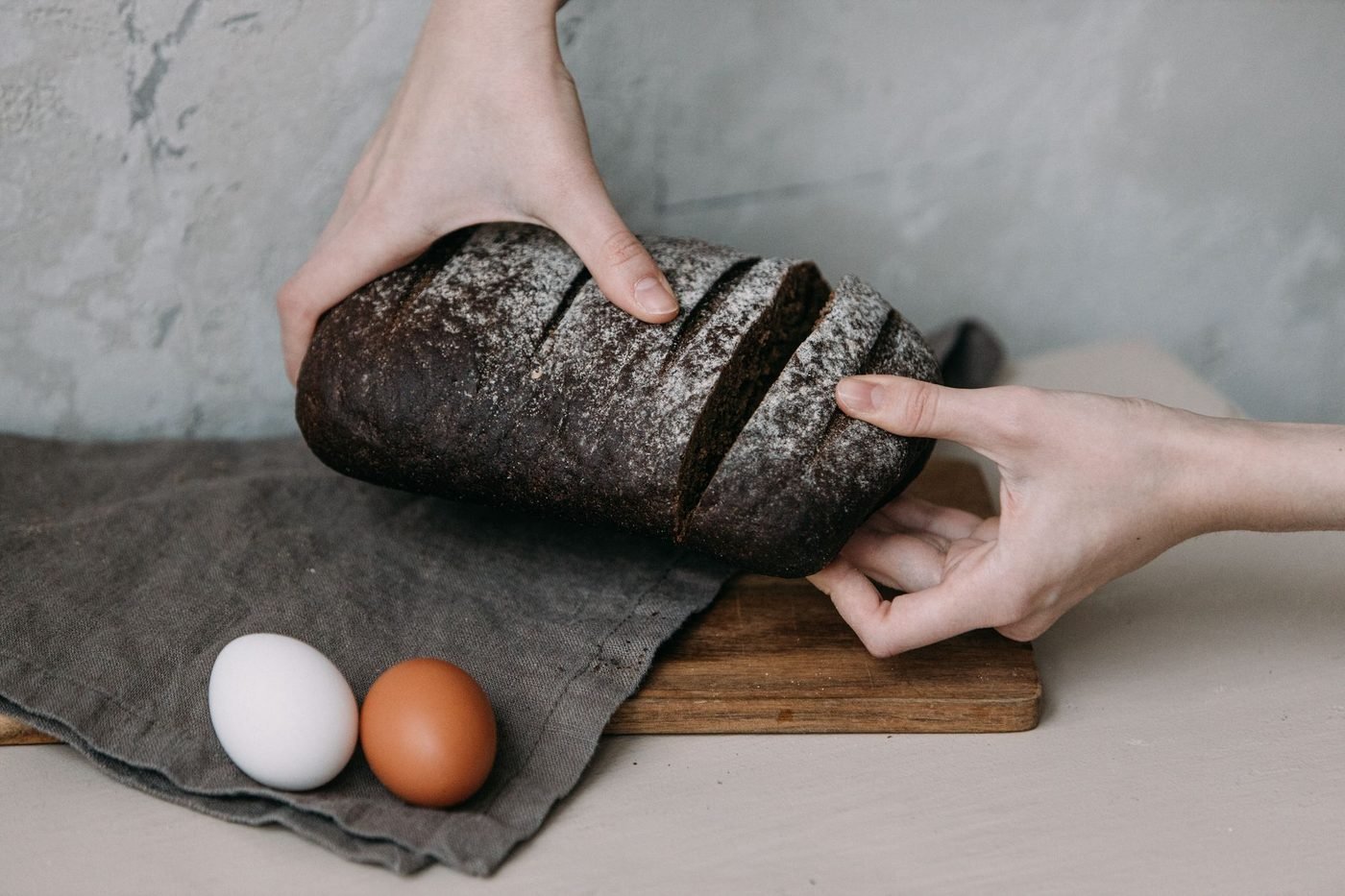 Hände halten ein angeschnittenes Roggenbrot über Leinentuch und Holzbrett